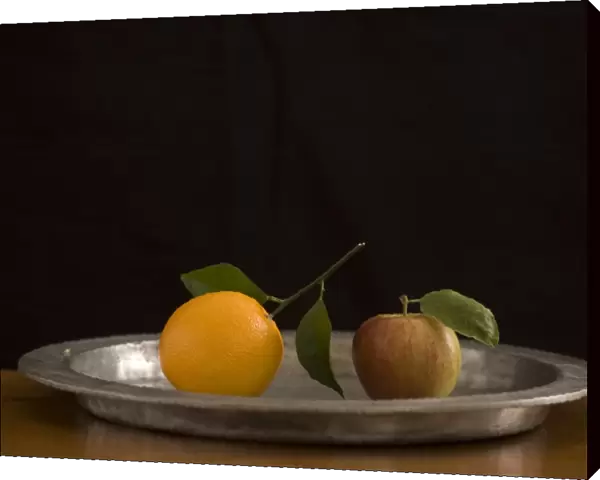 Apple and orange with leaves on pewter charger with black background credit: Marie-Louise