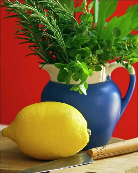 Lemon on chopping board with knife and bunch of mixed garden herbs in blue jug against