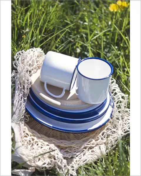 Enamel and paper cups and saucers piled up on a string bag on the grass - part of
