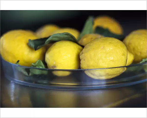 Natural, mediterranean lemons with leaves in glass dish on stainless steel counter