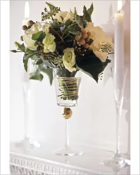 Arrangement of white roses and greenery in tall goblet glass between candles in tall