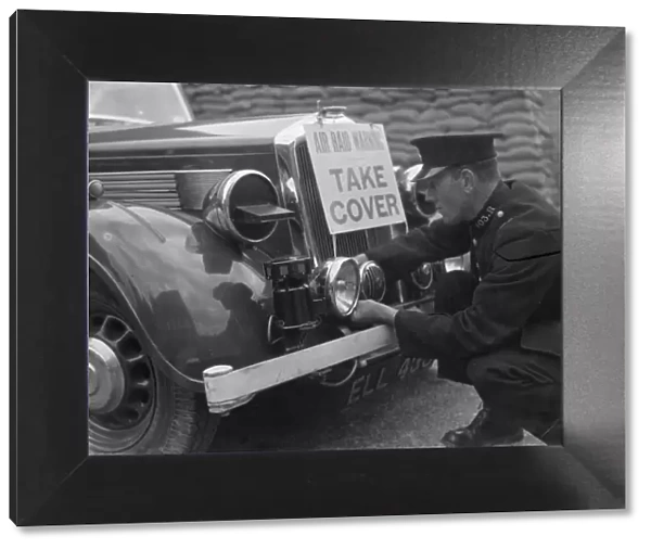 A policeman is fitting an air raid siren along with a warning sign to a police car