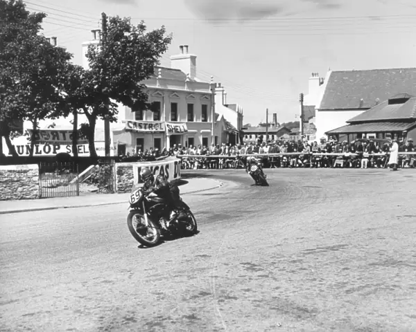 Competitors passing Ramsay Town Square at speed. Number 69 is W. J. Evans on AJS and number 90 is C