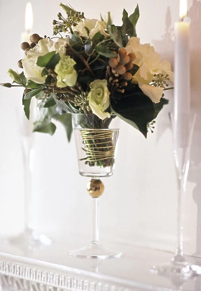 Arrangement of white roses and greenery in tall goblet glass between candles in tall