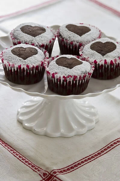Five chocolate muffins decorated with icing sugar in heart shape on white cake stand credit