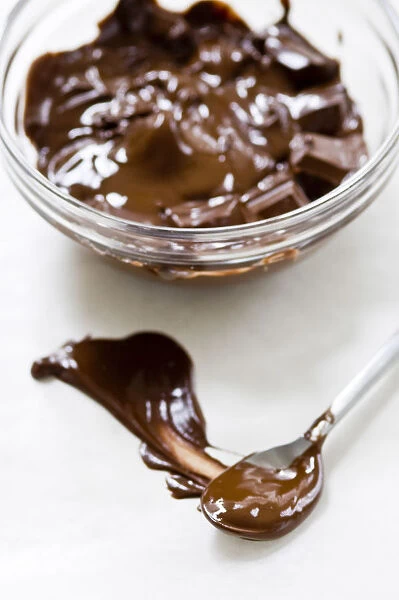 Glass bowl of melting chocolate with spoon smearing chocolate on white surface credit