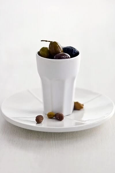 Mixed Italian olives in tall white ceramic pot on white plate with stones on white