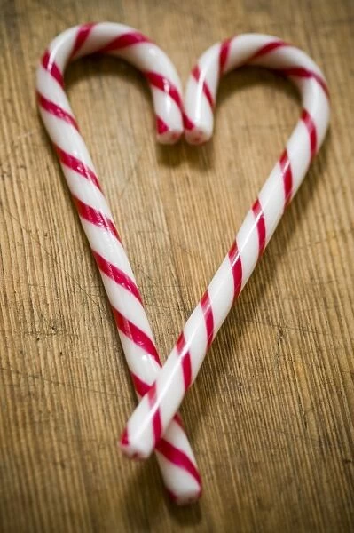 Two red and white striped candy canes on wooden surface crossed to make heart shape