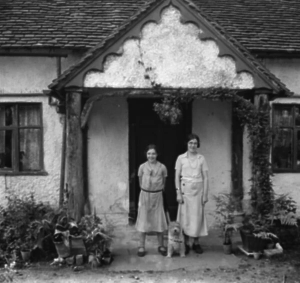 Two sisters and a dog pose in front of a cottage. 1936