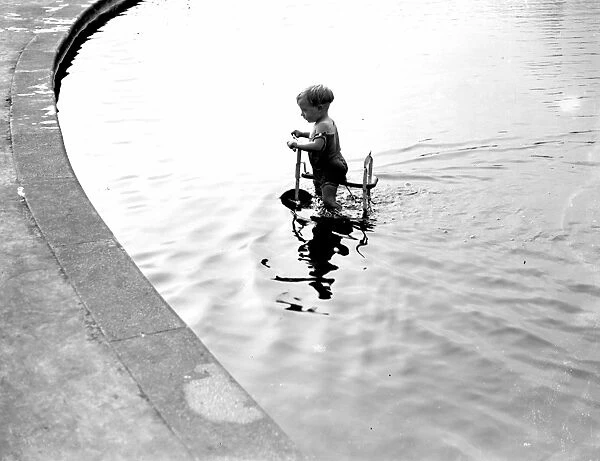 Toddler trying to ride his trike in the water. 1933