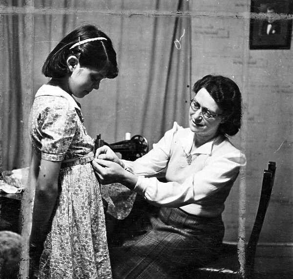 Woman sewing using a sewing machine and fitting a young girls dress [no caption