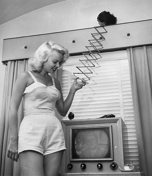 A woman uses an extendable duster whilst watching the television. 1950s ?TopFoto