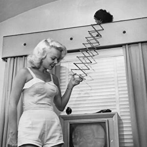 A woman uses an extendable duster whilst watching the television. 1950s ?TopFoto