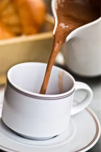 Pouring hot chocolate from jug into cup and saucer on cafe table, France, with brioche Pouring hot chocolate from jug into cup and saucer on cafe table, France, with brioche