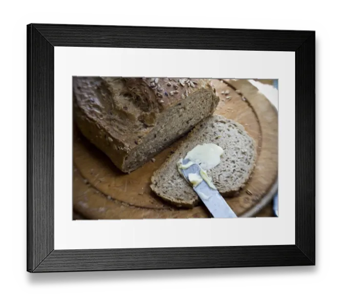 Framed Print : Buttering a slice of bread from a wholegrain, rye and walnut loaf on old wooden board