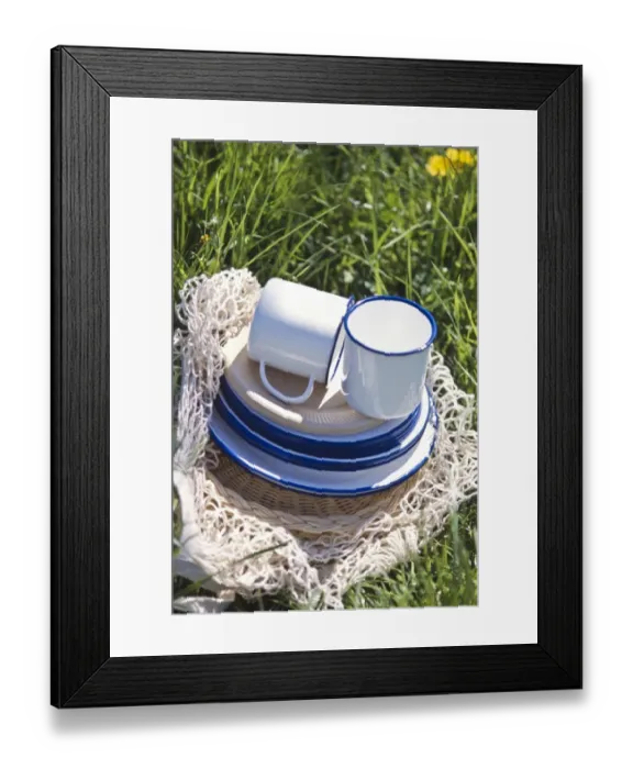 Framed Print : Enamel and paper cups and saucers piled up on a string bag on the grass - part of