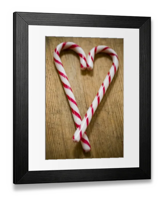 Framed Print : Two red and white striped candy canes on wooden surface crossed to make heart shape