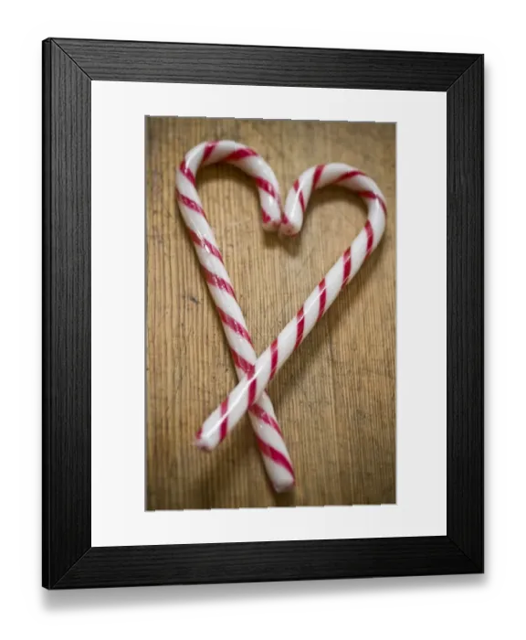 Framed Print : Two red and white striped candy canes on wooden surface crossed to make heart shape
