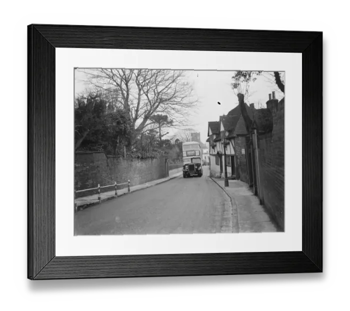 Framed Print : A double decker bus driving past some old cottages on a street in Ashford, Kent Framed Print : A double decker bus driving past some old cottages on a street in Ashford, Kent