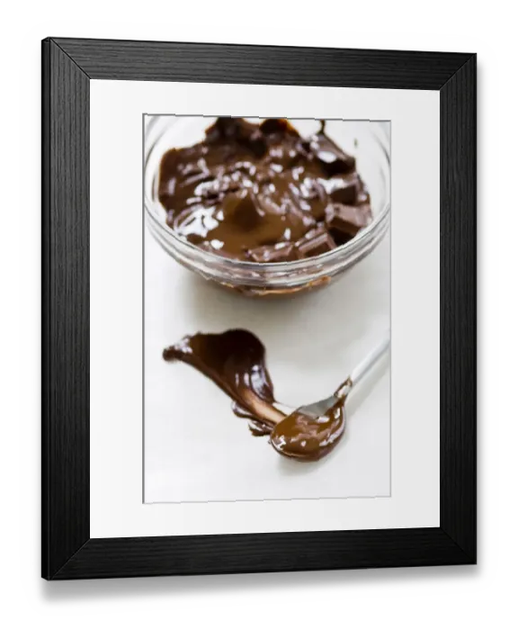 Framed Print : Glass bowl of melting chocolate with spoon smearing chocolate on white surface credit