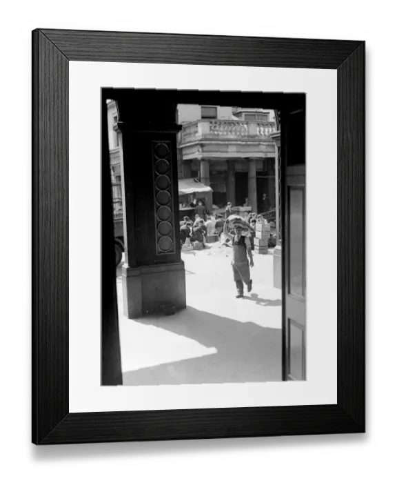Framed Print : A market porter transports a sack of vegetables on his head, while women market traders sit