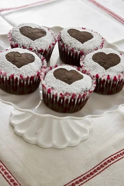 Five chocolate muffins decorated with icing sugar in heart shape on white cake stand credit