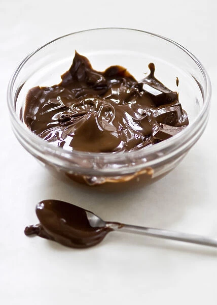 Glass bowl of melting chocolate with spoon on white surface credit: Marie-Louise
