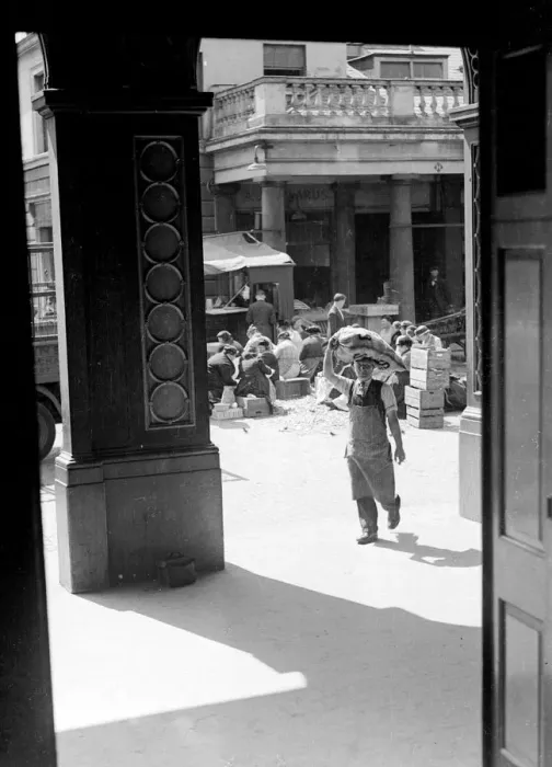 Framed Print : A market porter transports a sack of vegetables on his head, while women market traders sit