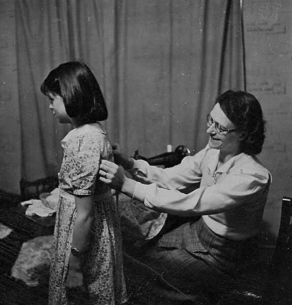 Woman sewing using a sewing machine and fitting a young girls dress [no caption