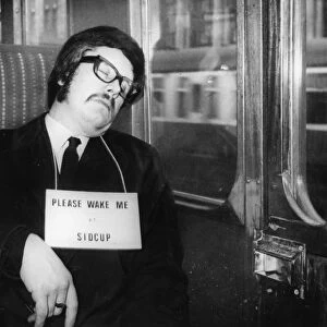 Commuter asleep on the train wearing a sign saying Please wake me up at Sidcup