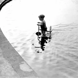 Toddler trying to ride his trike in the water. 1933