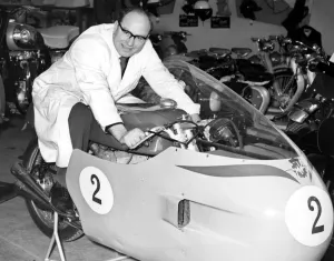Motorsport John Surtees Prints: A mechanic sits astride John Surteess racing MV Augusta motorbike inside the Surtees