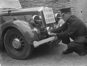 A policeman is fitting an air raid siren along with a warning sign to a police car
