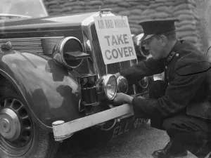 Preparation Prints: A policeman is fitting an air raid siren along with a warning sign to a police car