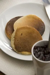 Three Scotch pancakes on a white plate with small pot of raspberry jam credit