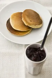 Three Scotch pancakes on a white plate with small pot of raspberry jam credit
