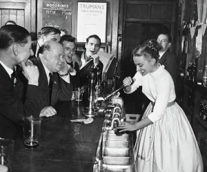 A woman barmaid pulls a pint of beer for male customers in a London pub, England. 1950s