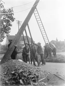 workers use ladders and poles erect an electric pylon next to a road in Trottiscliffe